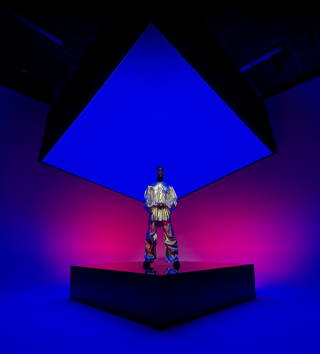 شخص يرتدي بدلة معدنية يقف على مسرح مربع في الوسط ويشمل الخلفية التي تعرض كاميرا Ultra Wide على iPhone 16 Pro Max